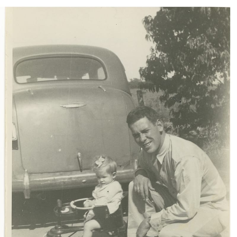 A man kneeling down next to a child in front of an old car.
